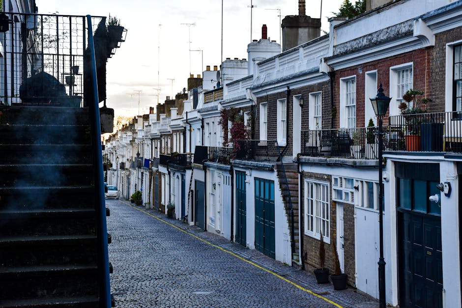 A narrow residential street with a row of terraced houses featuring brick and white-painted facades, each with small balconies and potted plants. The street is paved with cobblestones, and a black wro