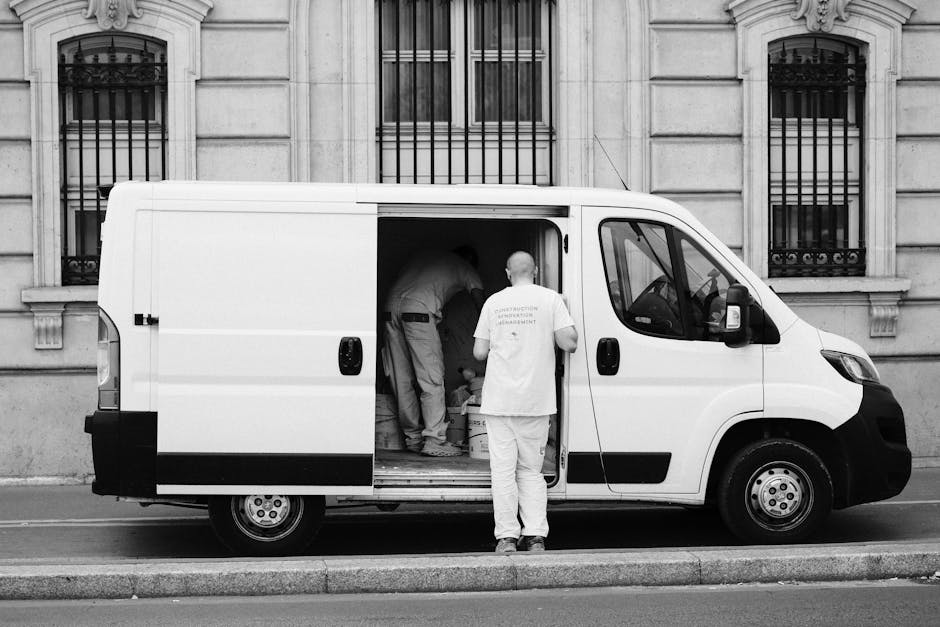 A white cargo van parked on a city street beside a tall, light-colored building with multiple windows and architectural details. The van's rear double doors are open, revealing various packing materia